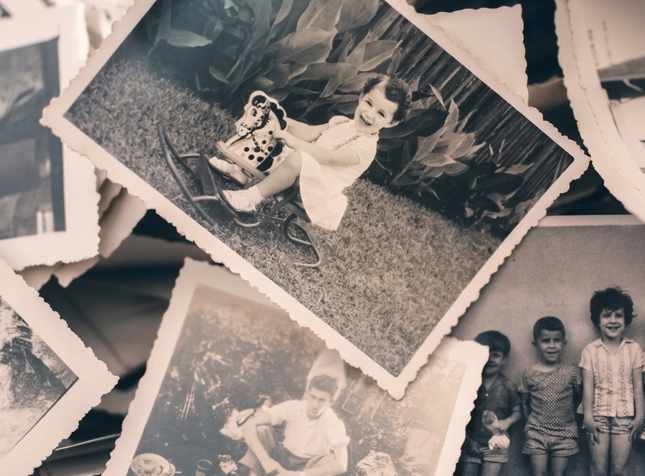 En stämningsfull närbild på en samling gamla vintage-fotografier med tandade, vågiga kanter spridda i en hög. Bilderna föreställer olika scener som ett barn på en gungbåt, en grupp barn och en man, och symboliserar minnen och livshistoria.