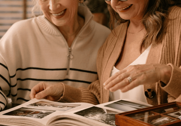 Två äldre kvinnor ler och bläddrar i ett fotoalbum fyllt med gamla bilder, en illustration av att börja skriva en självbiografi baserad på personliga minnen.
